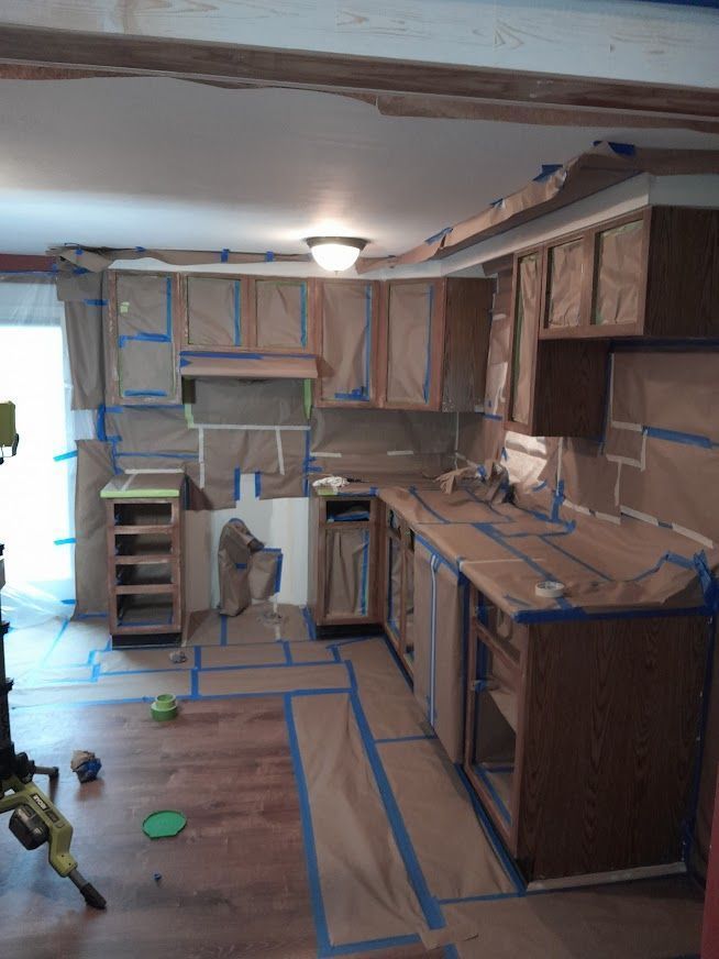 Kitchen cabinets and surfaces covered in brown paper and blue painter's tape, preparing for painting.