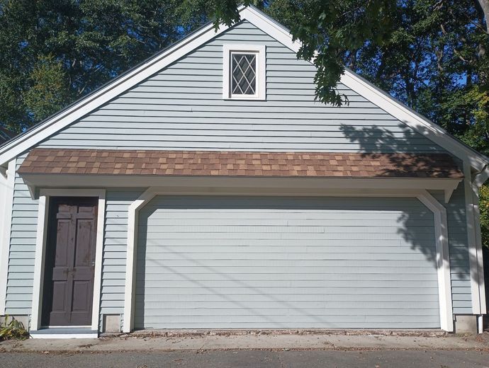 Garage with light blue siding, brown roof, and dark brown door. A small window is at the peak.