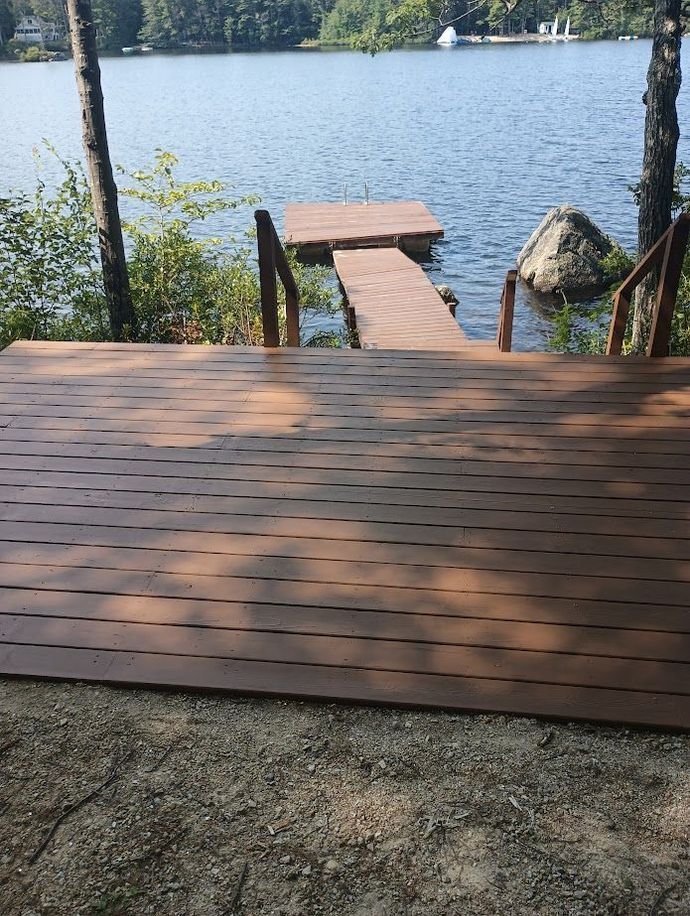 Wooden deck and dock leading to a lake, brown wood. Sunlight, trees, and blue water.