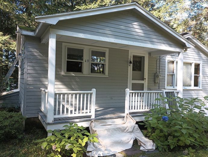 Small, gray house with a porch and white railings; overgrown shrubs in front.