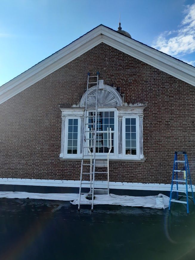 Brick building facade with a window, white trim, ladder, and blue ladder against a blue sky.