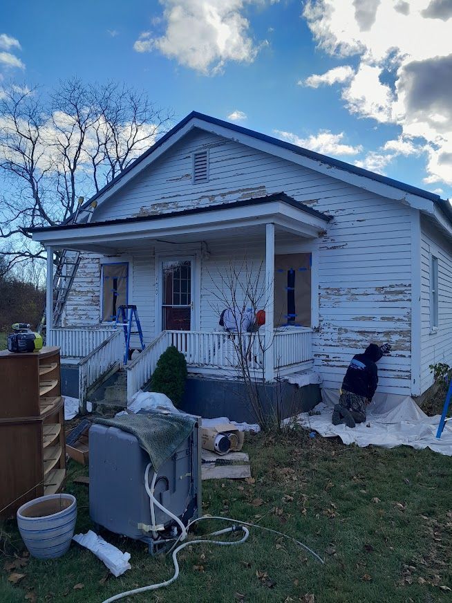 House exterior with peeling white paint, porch, and a person working on the side.  Outdoor setting with cloudy sky.