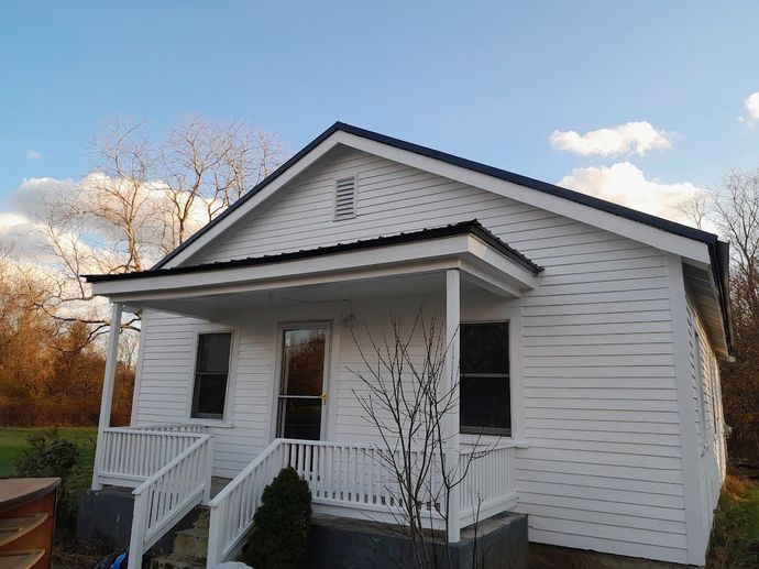 White house with black roof and small porch, set against a cloudy blue sky.