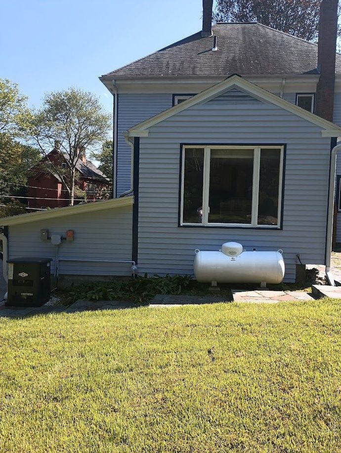 Back of a light blue house with a white propane tank and a large window. Green lawn.