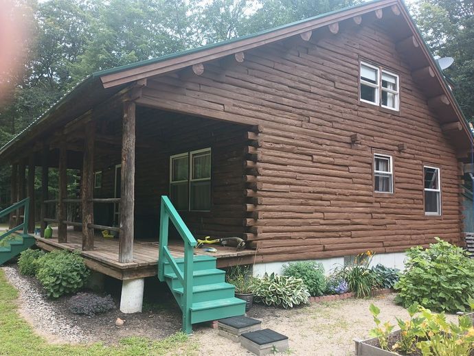 Brown log cabin with green porch and stairs. White-framed windows, surrounded by landscaping.