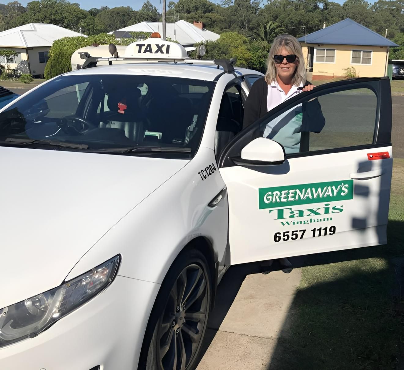 Woman standing by a white taxi, door open. Greenaway's Taxis sign on the side, phone number — Greenaway's Taxi in Wingham, NSW