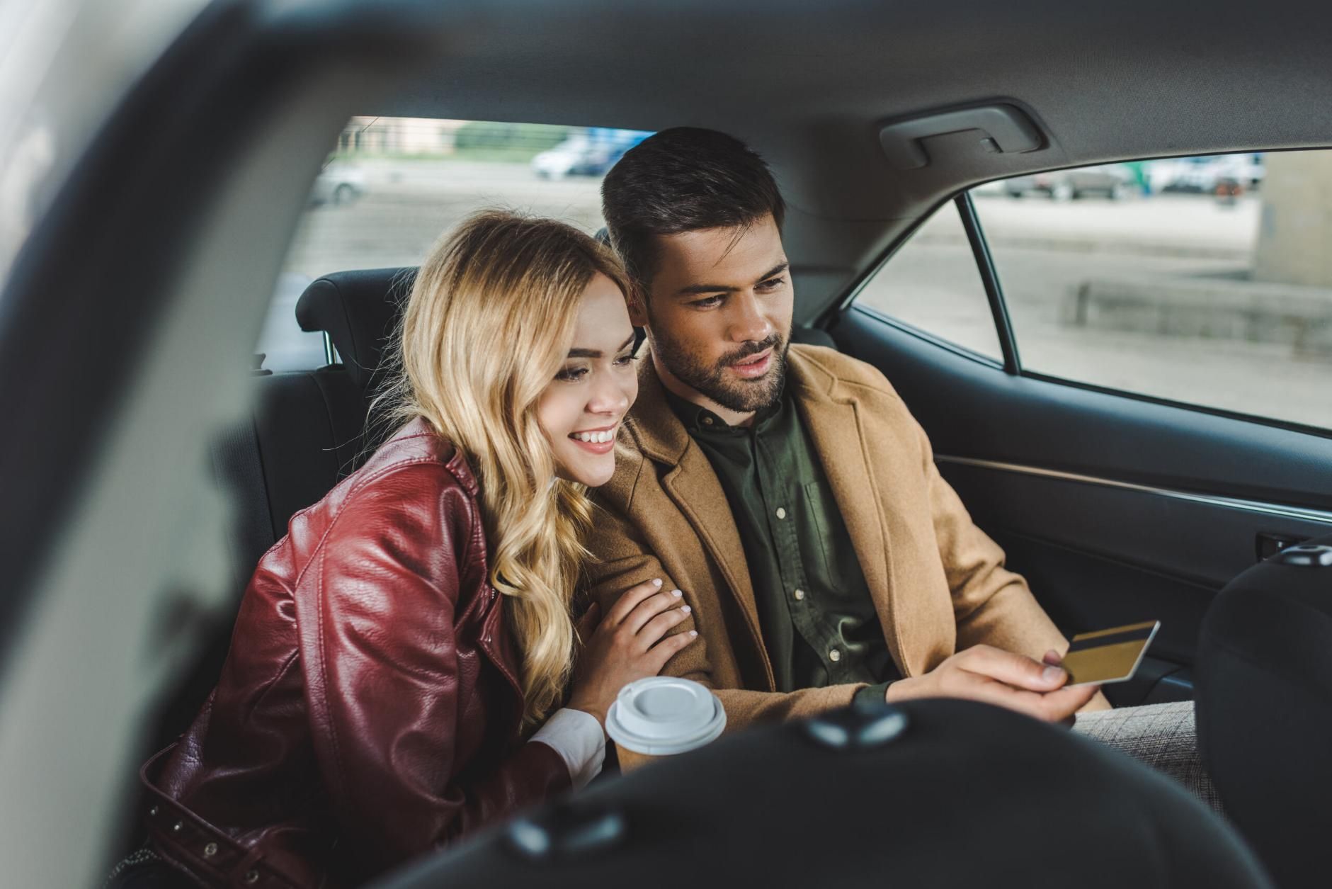 Couple in the backseat of a car looking at a credit card, smiling. Beige and red clothing — Greenaway's Taxi in Cundletown, NSW