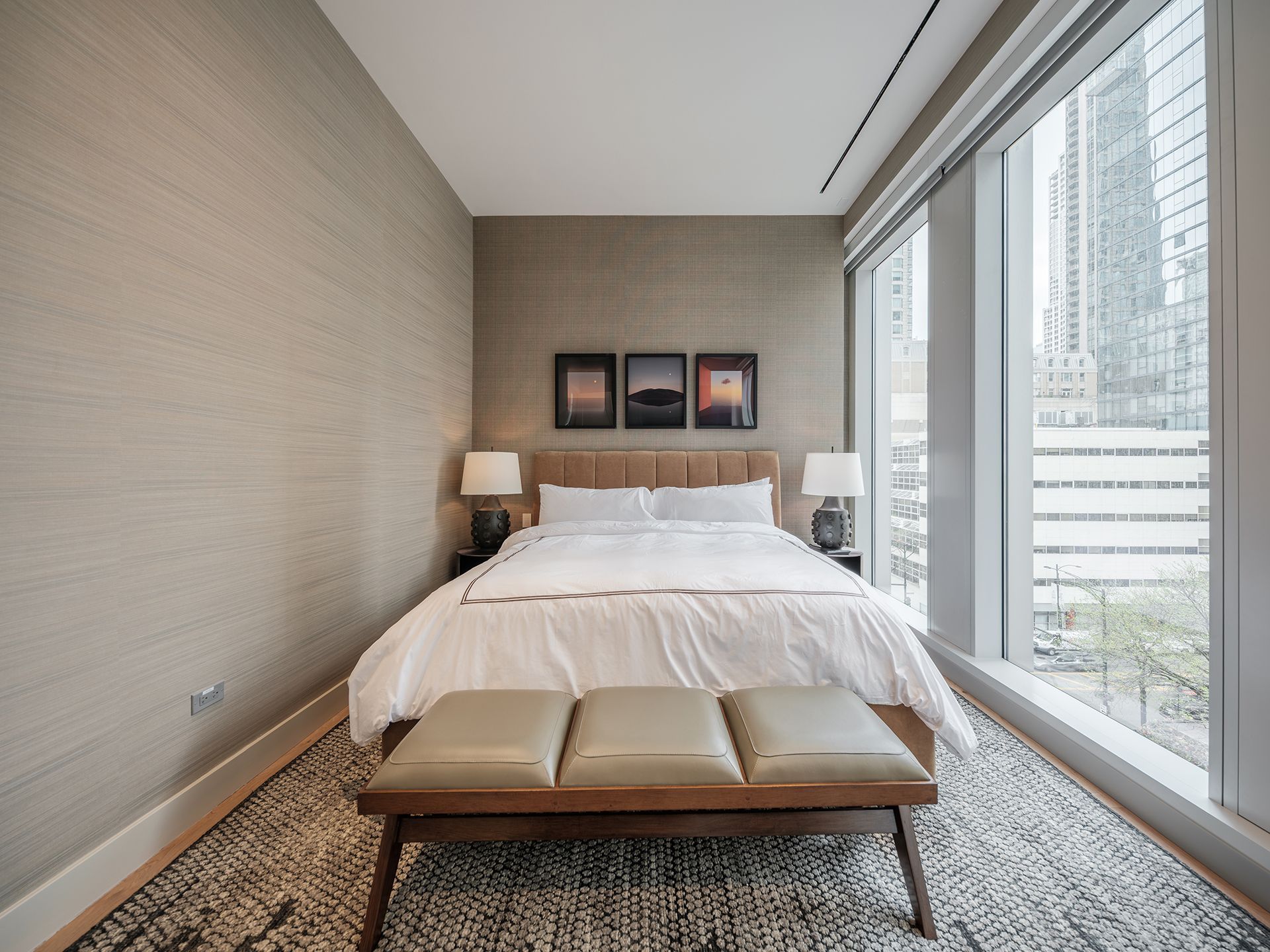 A bedroom with a bed , bench , lamps and a large window.