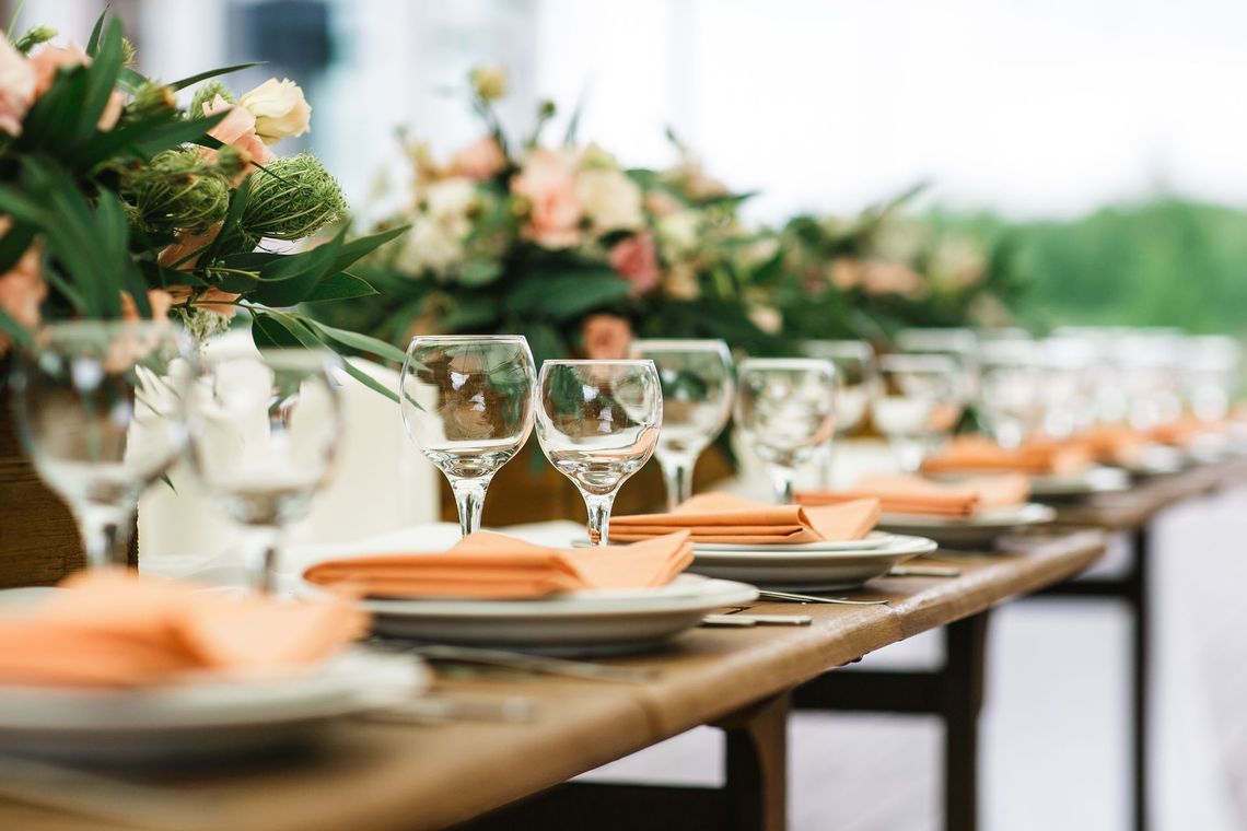 A long wooden table set for a meal with floral arrangements, plates, glasses, and orange napkins.