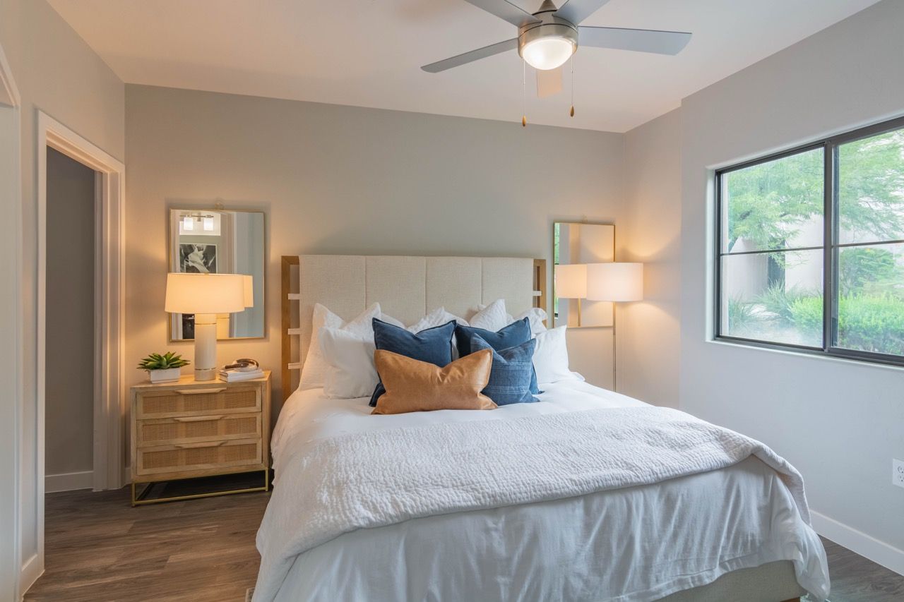 Bedroom with a padded headboard bed, white bedding, blue and orange pillows, two nightstands, lamps, and a window at Legacy Apartments at Dove Mountain in Marana, AZ.