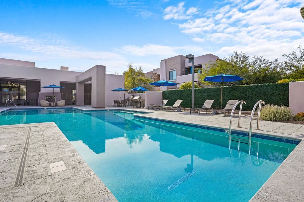 Outdoor apartment pool with blue umbrellas, lounge chairs, and a paved deck.