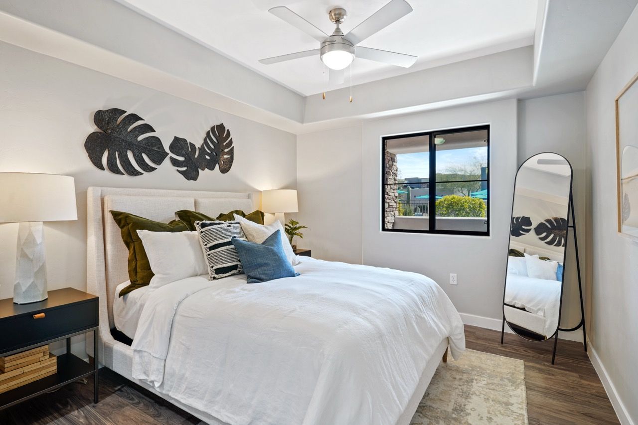 Bedroom in an apartment with white bedding, nightstands, lamps, a window, and a full-length mirror at Legacy Apartments at Dove Mountain in Marana, AZ.