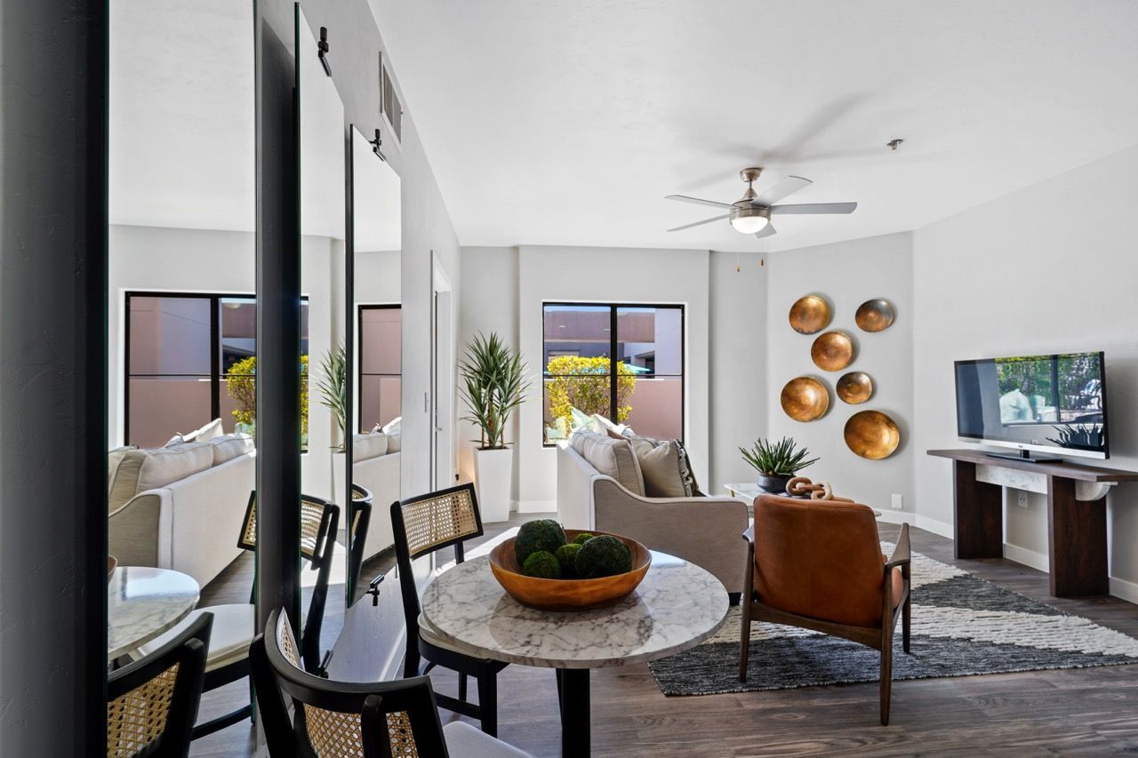 Living room in a modern apartment with a round marble dining table, seating, and a TV near large windows at Legacy Apartments at Dove Mountain in Marana, AZ.