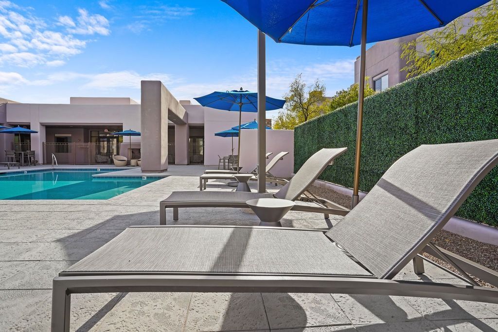 Outdoor pool area with lounge chairs, blue umbrellas, and a green hedge at Legacy Apartments at Dove Mountain in Marana, AZ.