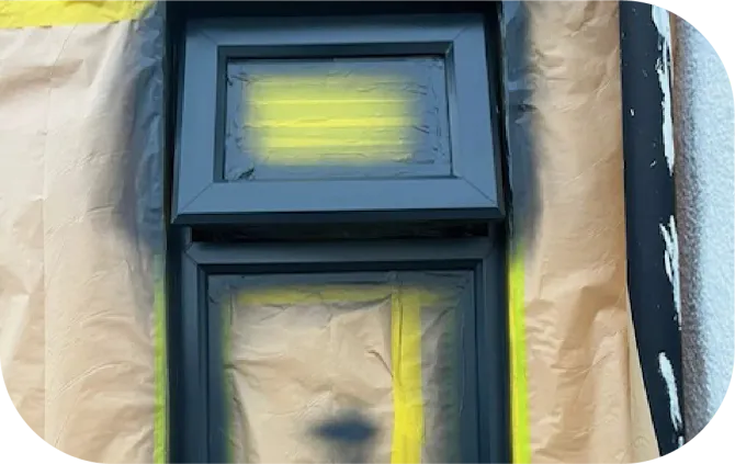 A window frame being spray-painted black, with masking paper protected by yellow tape surrounding it.