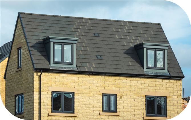 A tan stone house with a dark gray shingled roof, featuring two dormer windows and several smaller windows.
