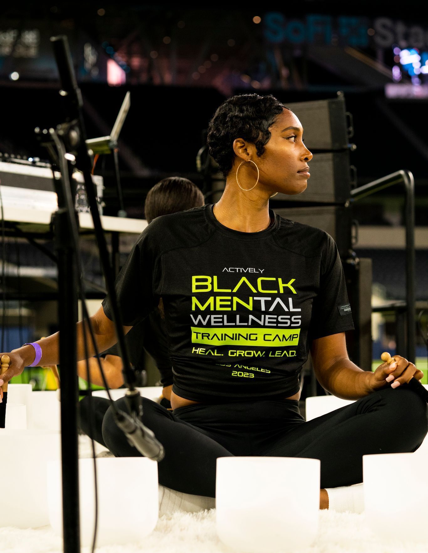 A woman wearing an Actively Black black mental wellness t-shirt is sitting on a stage playing sound bowls.