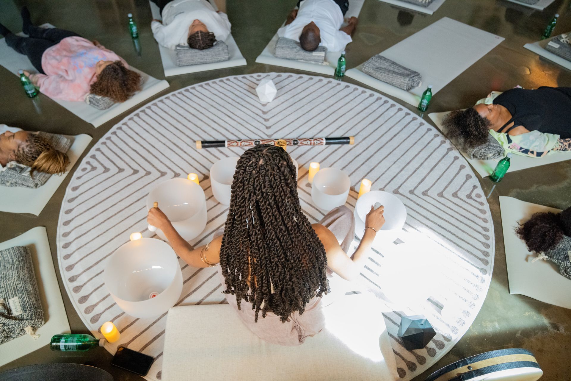 A woman is sitting in a circle of people laying on yoga mats while playing sound bowls.