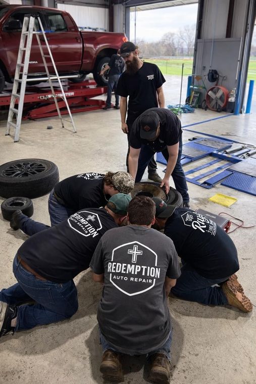 People kneeling around a car tire in a garage, working together with tools and equipment nearby.