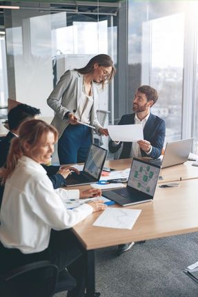 Colleagues in a glass-walled office meeting around laptops and papers, one person standing and discussing a document