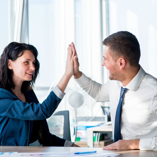 Two coworkers high-five across a desk in a bright office.