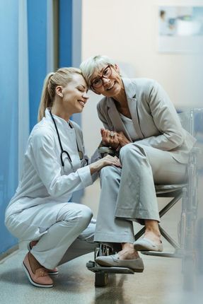 Doctor comforting elderly patient in clinic hallway, seated on exam bench and holding hands