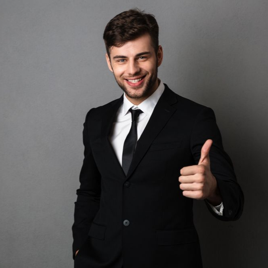 Smiling man in a black suit giving a thumbs-up against a gray background