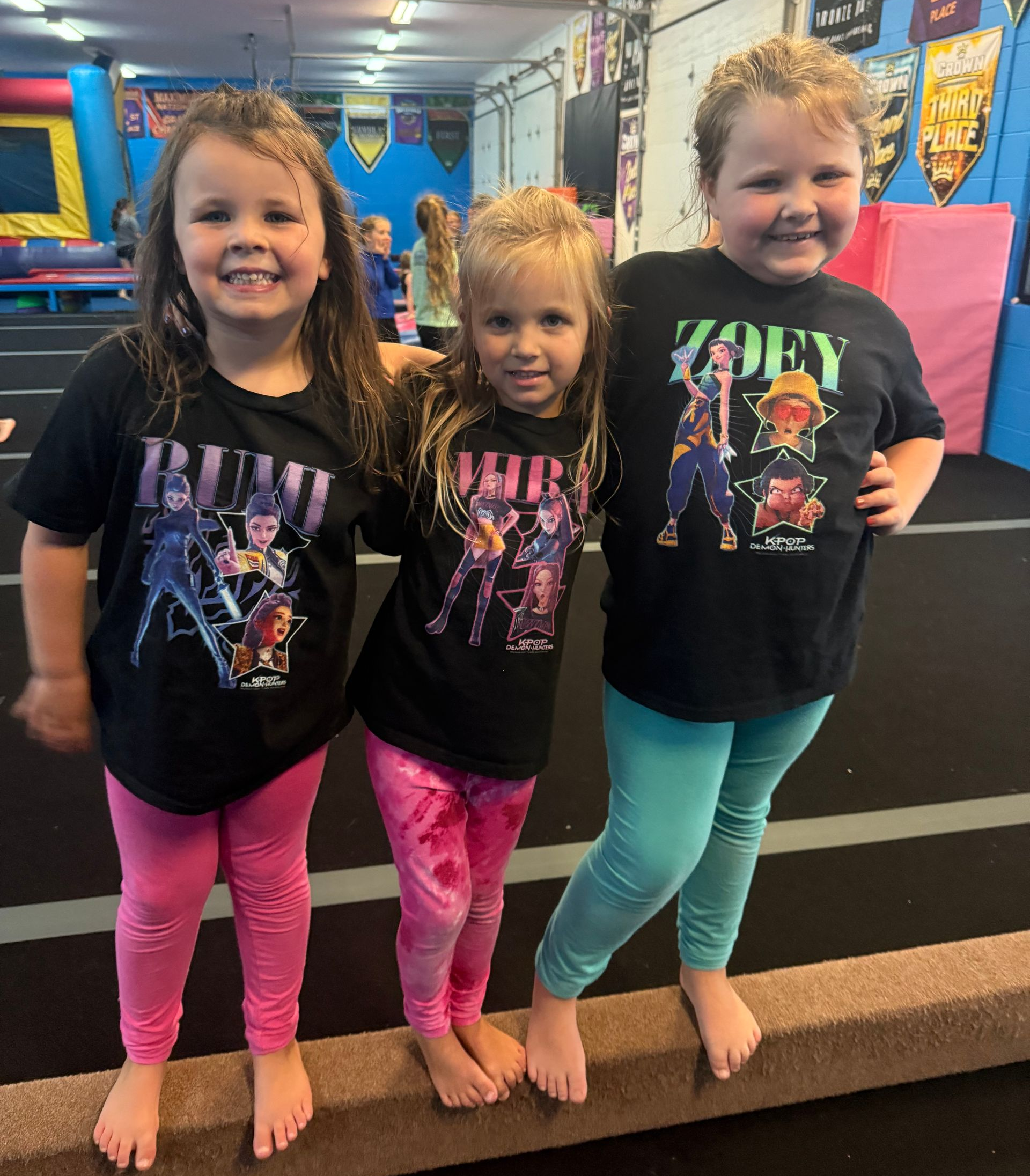 Three children standing in a gym, wearing black graphic t-shirts and colorful leggings, smiling at the camera.
