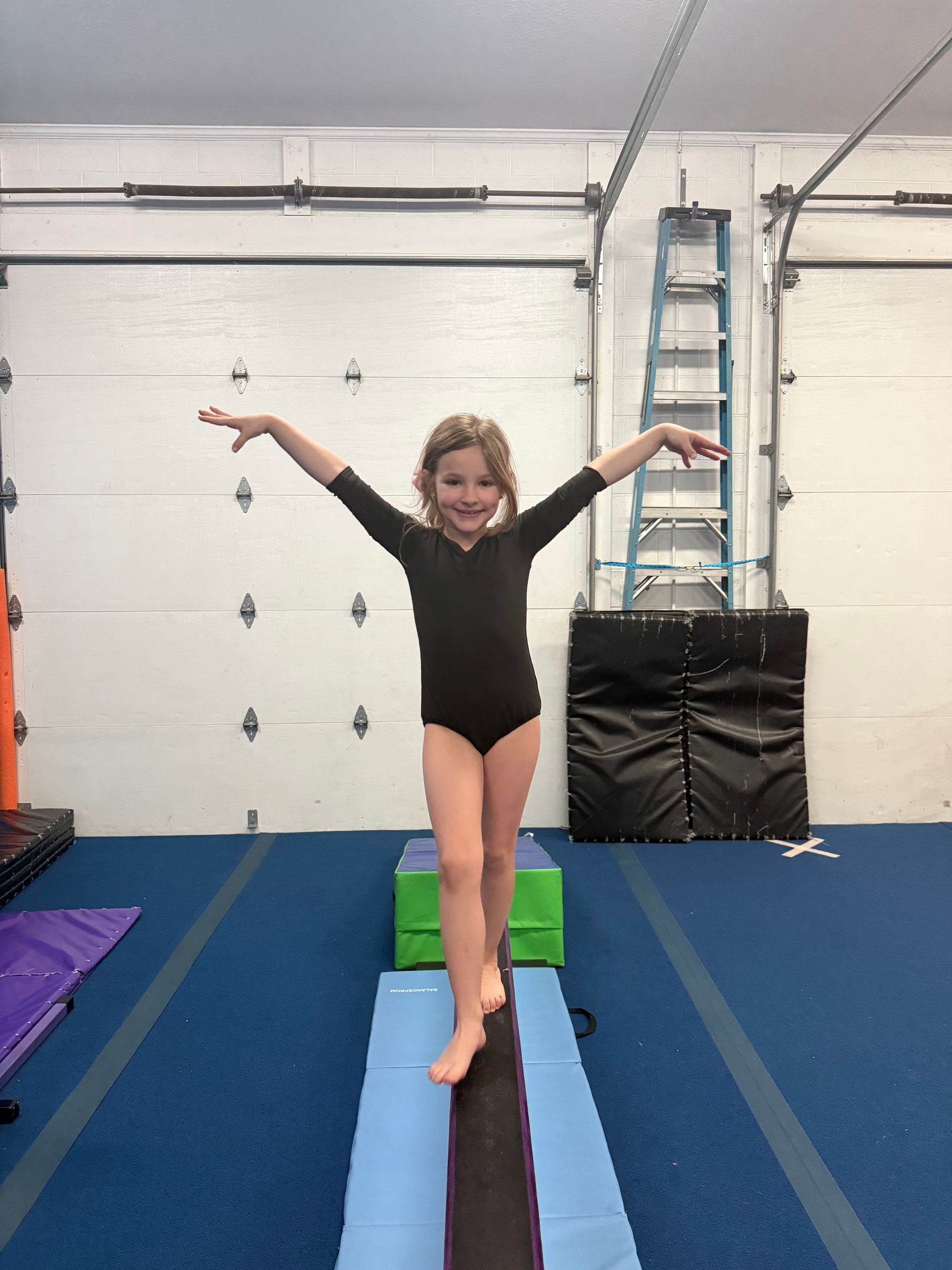 A child in a black leotard balances on a floor balance beam in a gym with arms extended.