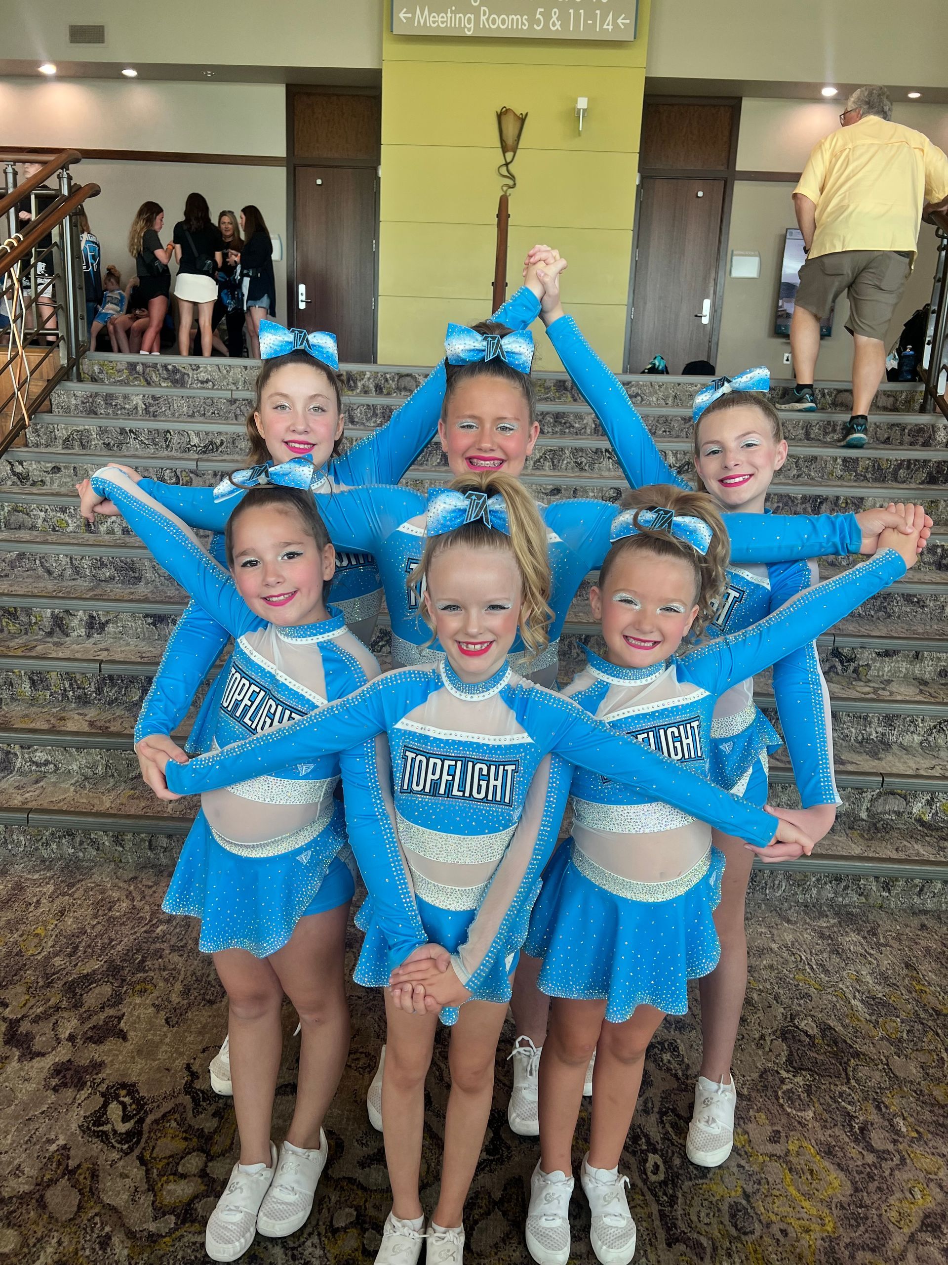 Six girls in matching bright blue cheerleading uniforms stand on stairs, smiling and linking arms in a star formation.