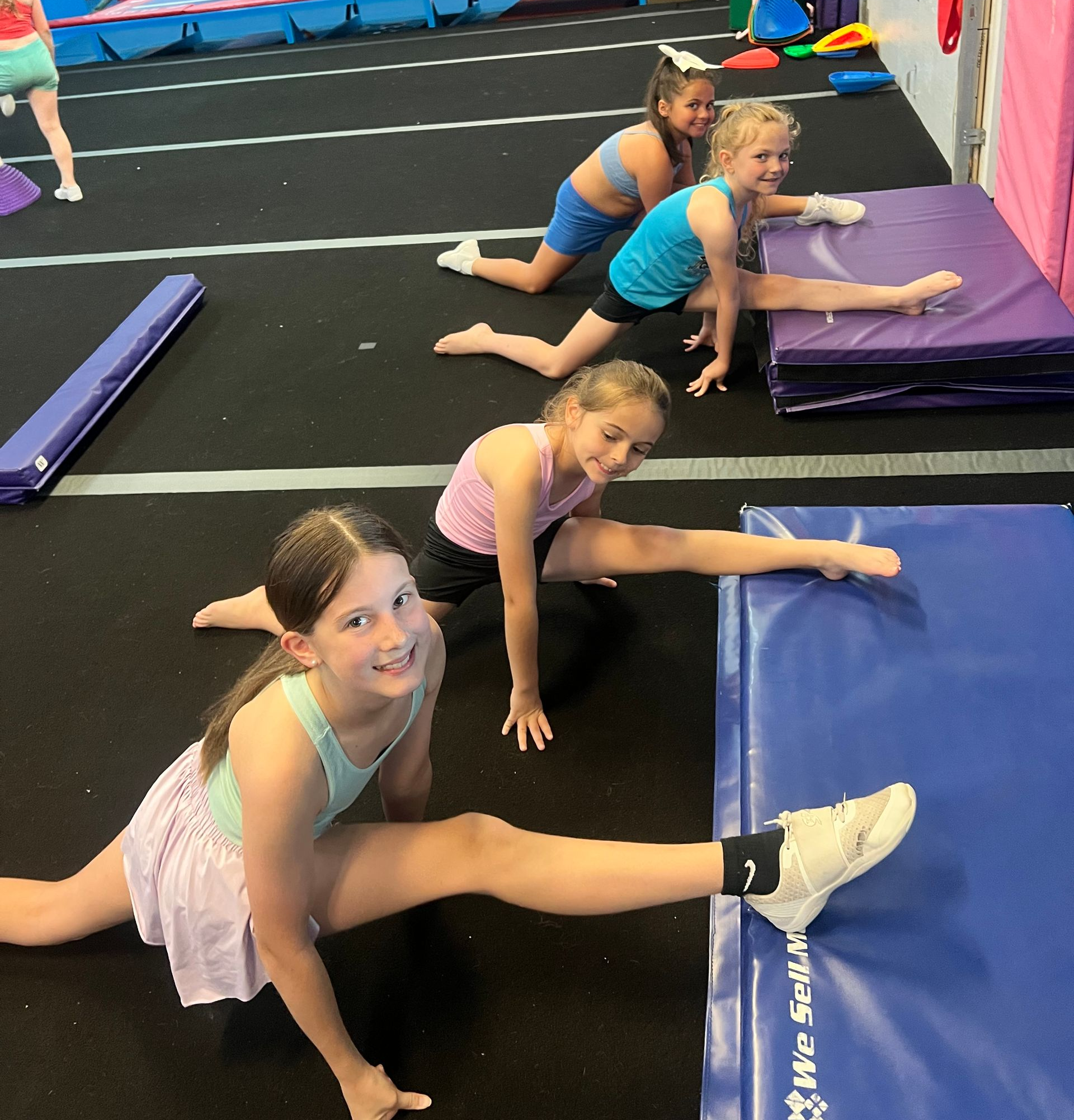 Four people in activewear stretch into splits on mats in a gym, with the closest person smiling at the camera.