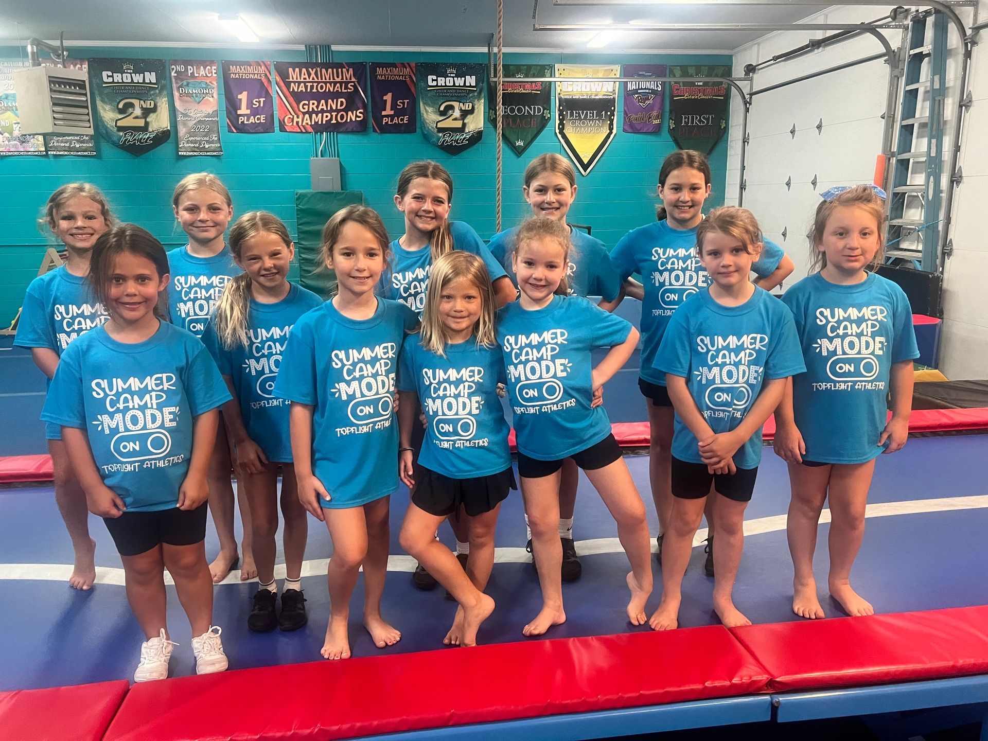 A group of children in matching blue t-shirts stand in a line at a gymnastics facility, smiling for the camera.