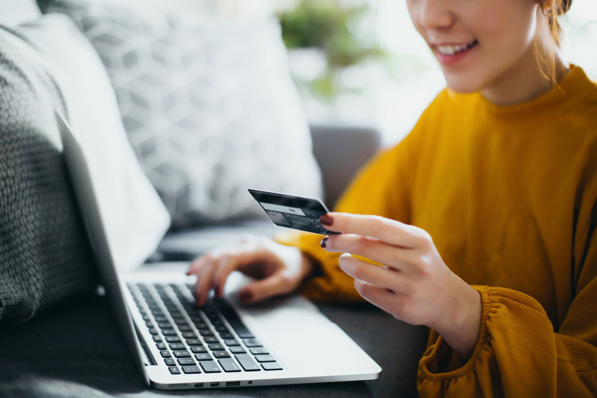 Woman smiles while holding a credit card and using a laptop, possibly shopping online. She's indoors on a sofa, wearing a yellow shirt.