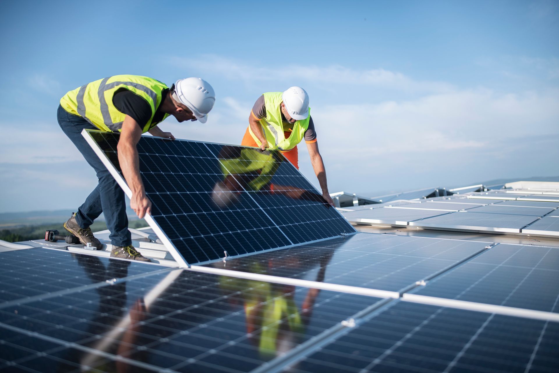 Two workers installing solar panels on a rooftop. They wear safety vests and helmets, working under a blue sky.