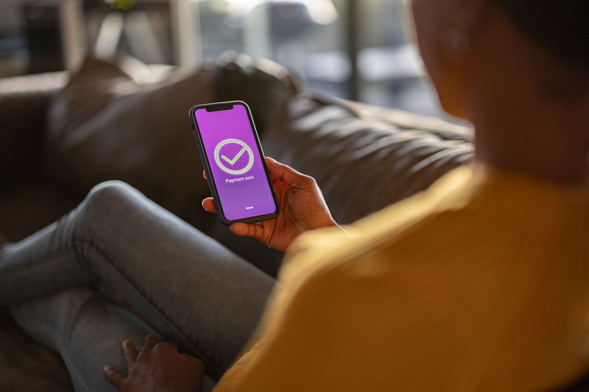 Woman sitting on a couch, holding a phone with a purple screen showing a check mark, indicating successful transaction.