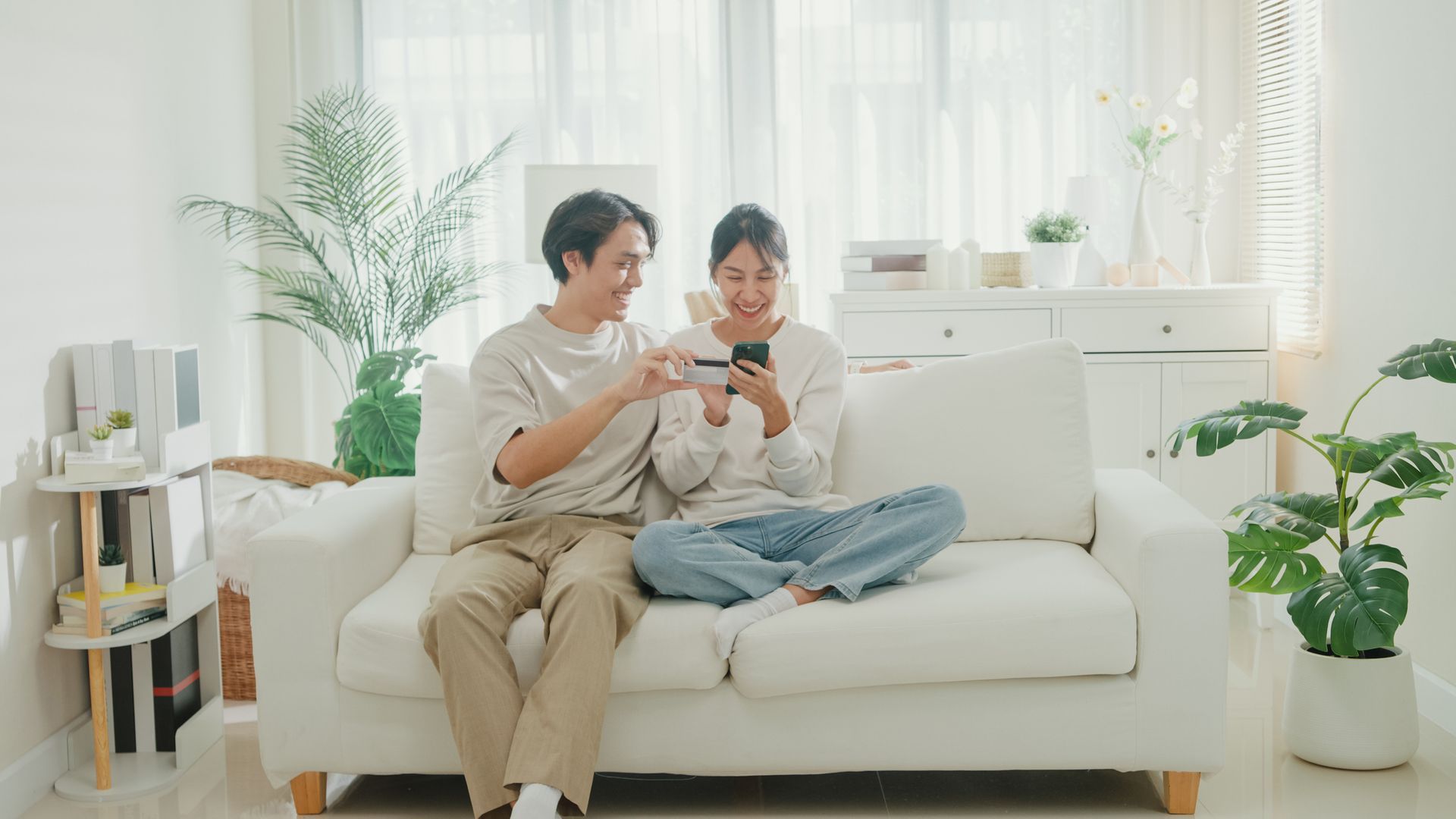 Young Asian couple on a white couch smiling at a phone. Man points, woman holds phone, bright living room with plants.
