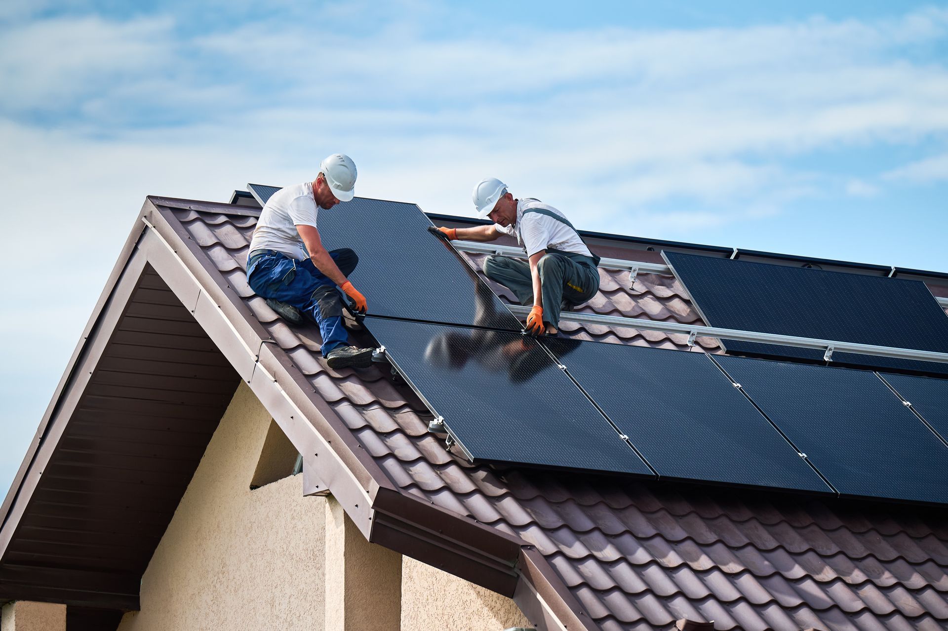 Two workers installing solar panels on a brown shingled roof against a cloudy sky.