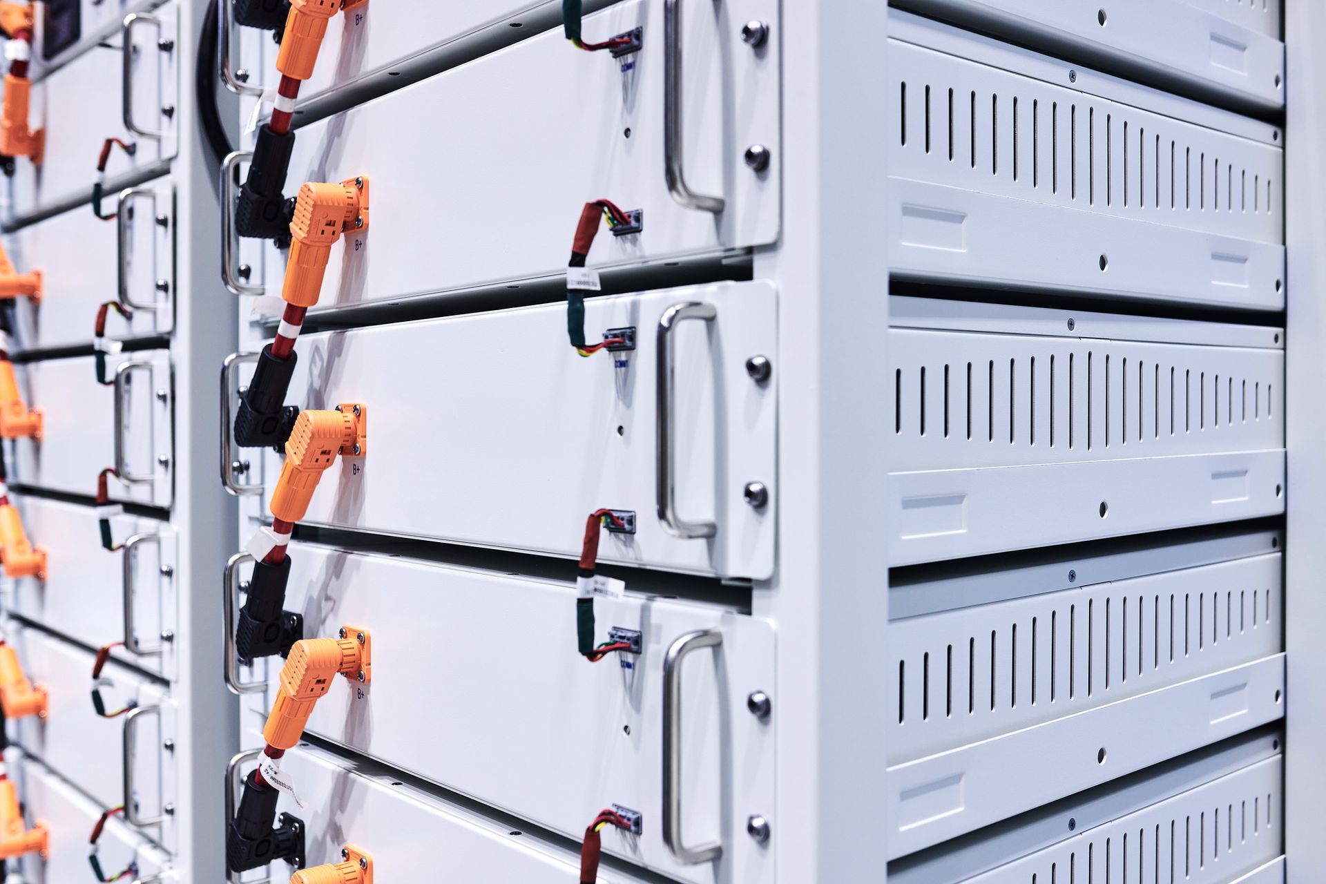 White battery storage units in a row with orange and black connectors. They are stacked in a room.