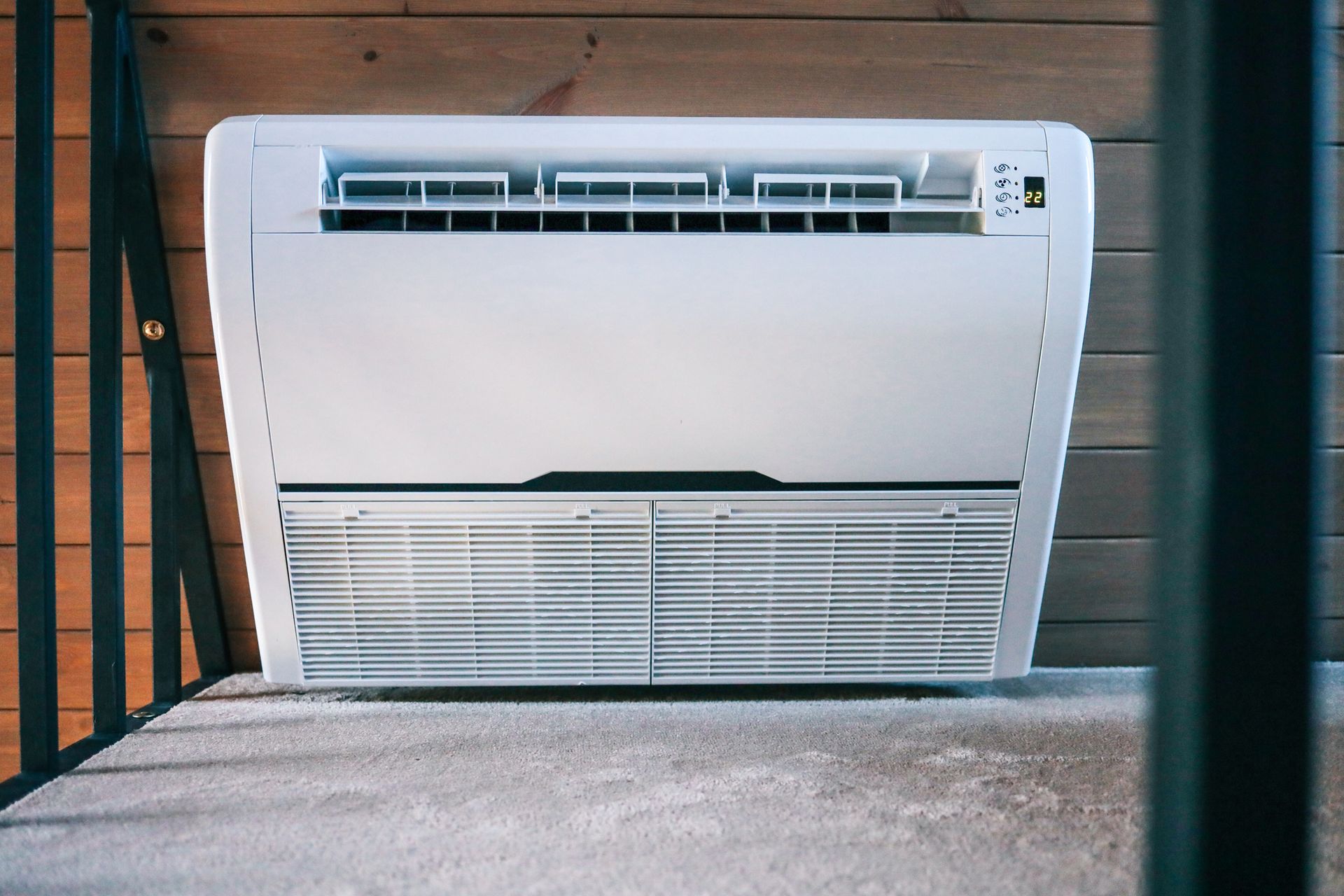 White wall-mounted air conditioner on a platform, with wooden paneling in the background. A black railing is partially visible on the left.