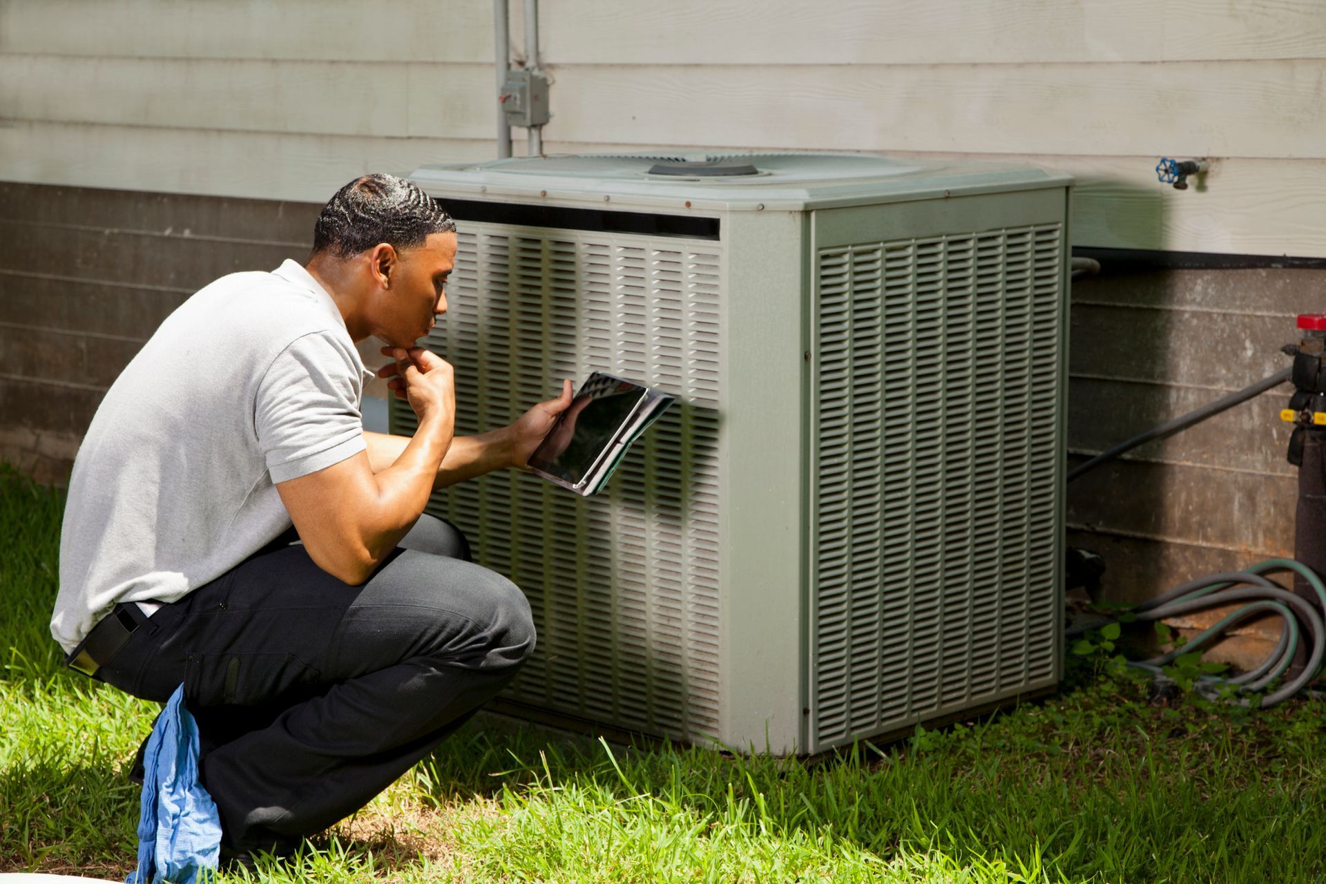 A man in black pants and a gray shirt inspects an air conditioning unit, looking at a tablet outdoors.