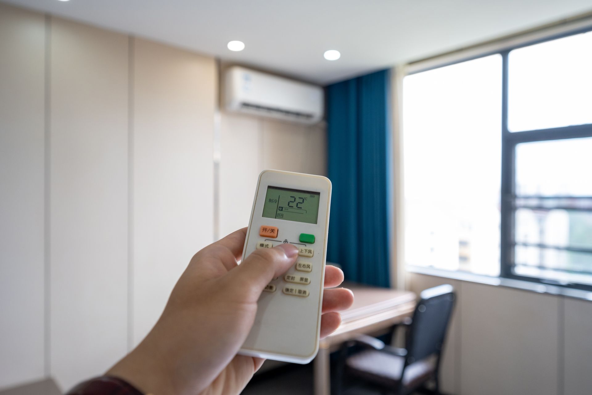 A person holds a remote control pointed towards a wall-mounted air conditioner in an office setting. The remote displays a temperature of 22.