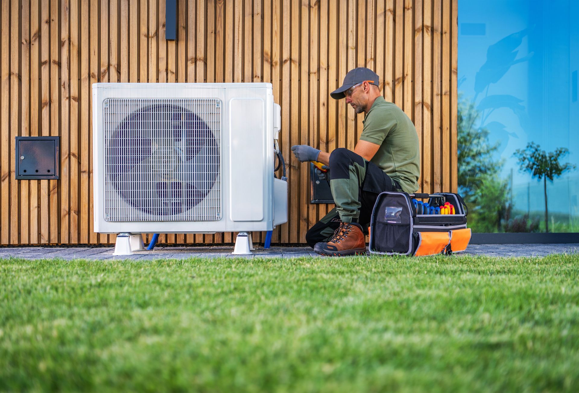 Man in work clothes repairs a white heat pump unit outdoors. The pump sits on a grassy lawn near a wooden-paneled wall and a glass window.