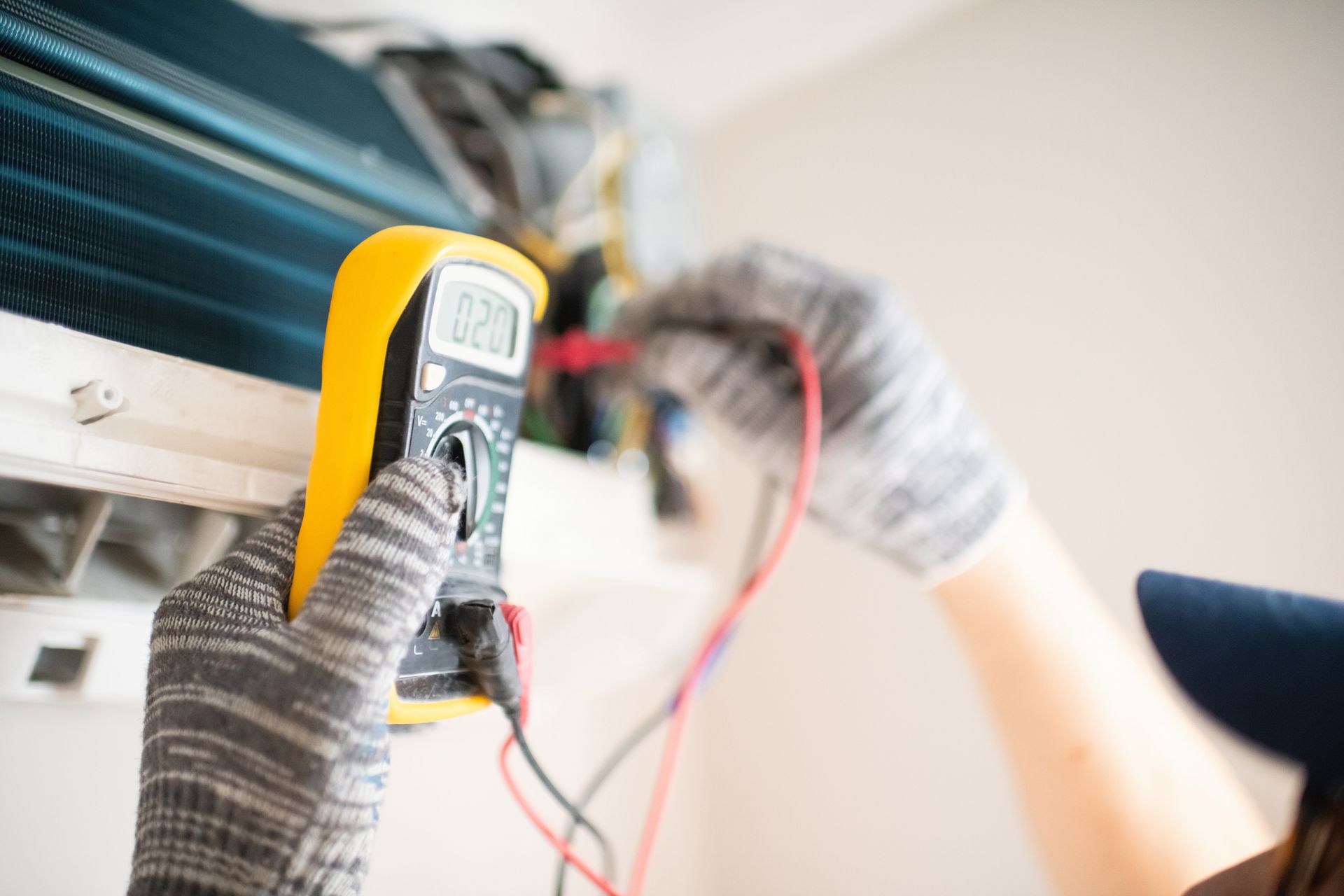 A person uses a multimeter to check an air conditioning unit's electrical components. They wear gloves and a cap while working indoors.