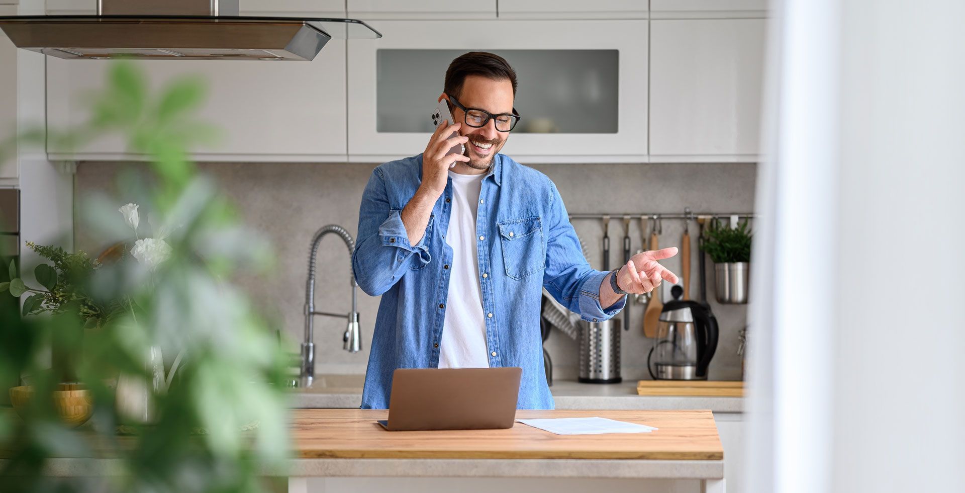 Man in denim shirt on a phone call in a kitchen, looking at a laptop and gesturing.