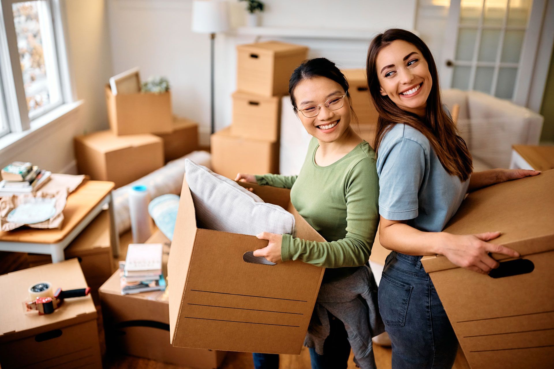Two women smiling, packing boxes in a room filled with moving boxes.
