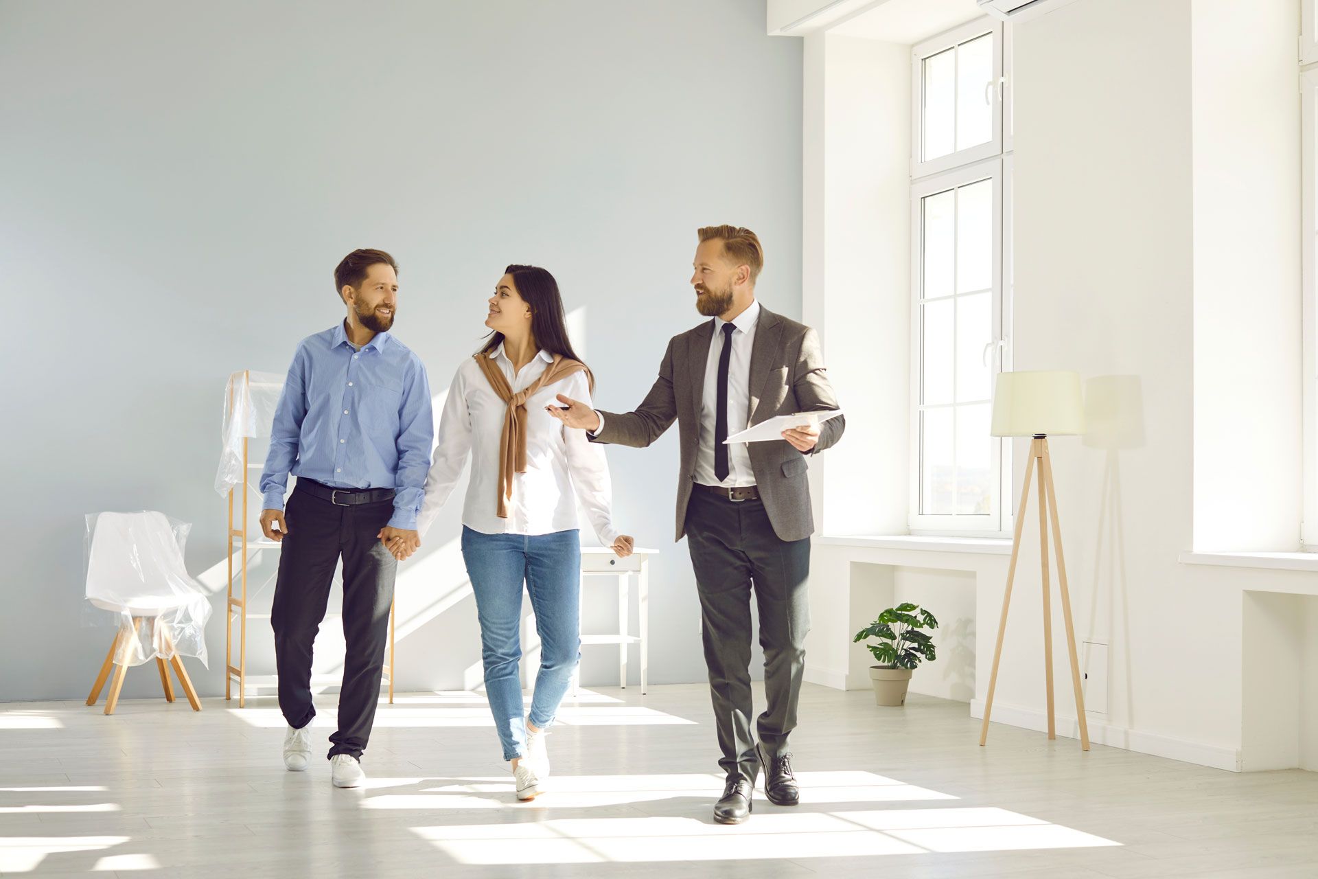 Real estate agent leading a couple through a bright, empty room with a large window.