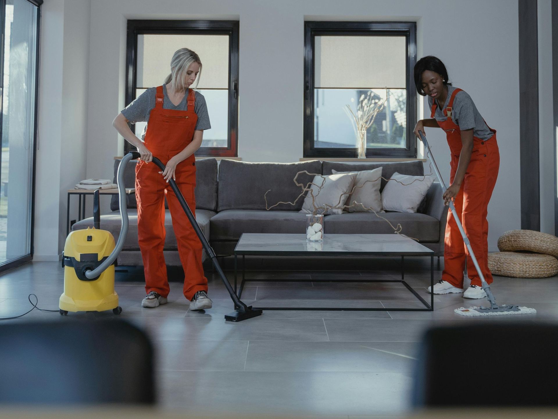 Two women are cleaning a living room with a vacuum cleaner and mop.