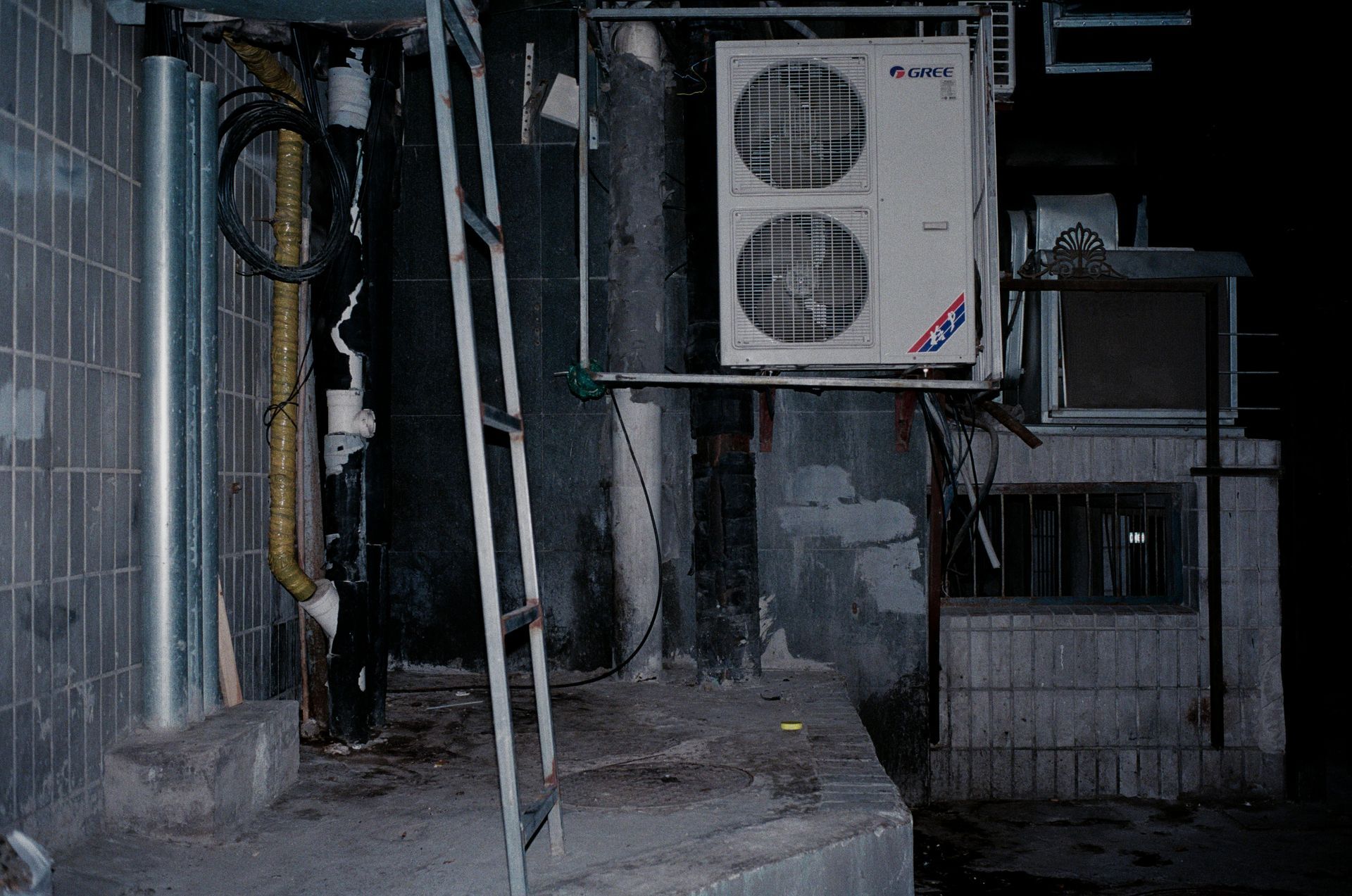 Dim alley with a wall-mounted air conditioner unit and pipes beside a concrete wall at night.