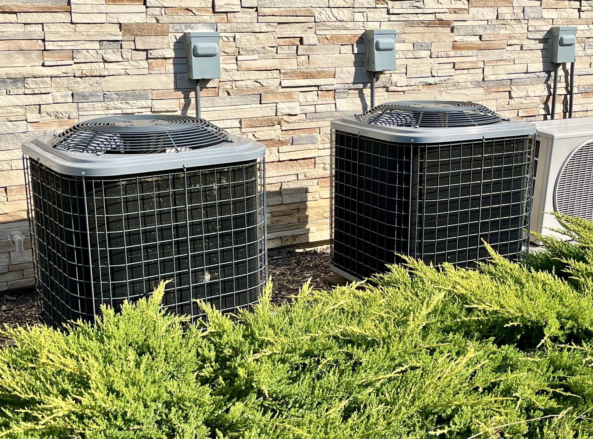 Two outdoor air conditioning units beside a stone wall and green shrubs