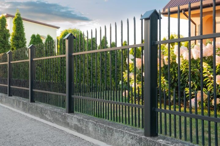 Black metal fence along a concrete curb, with a hedge and a house in the background.